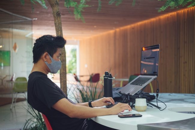 paul-hanaoka-OTqCo3fdWyw-unsplash work from home, young man with protective mask on his face working remotely with his laptop